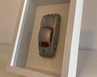 May include: A silver model car in a white frame. The car is a classic sports car with a rounded body and a black interior. The frame is made of wood and has a glass front.