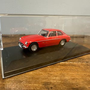 Puede incluir: Un coche modelo rojo dentro de una vitrina transparente. El coche en miniatura tiene ruedas plateadas y un exterior detallado. La vitrina se encuentra sobre una base negra, y el fondo es una superficie de madera.