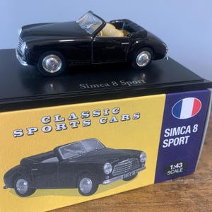 May include: A black Simca 8 Sport model car with a tan interior sits on a black display stand. The model is on a box labeled "Classic Sports Cars" and "Simca 8 Sport". The box has a French flag and a 1:43 scale indicator.