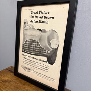 May include: Framed vintage advertisement for David Brown Aston Martin, featuring a classic sports car. The black and white print includes text about a hill climb record and the car's performance.
