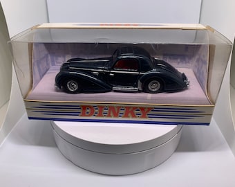 May include: A dark blue Dinky toy car in a clear plastic display case. The car is a vintage model with a red interior and chrome accents. The box has a white background with blue and gold stripes and the word "DINKY" in red letters.