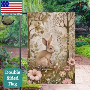 May include: A double-sided garden flag featuring a brown rabbit sitting in a field of pink and white flowers. The background is a soft beige with brown branches and leaves.