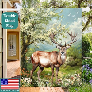 May include: A double-sided garden flag featuring a brown deer standing in a field of wildflowers. The deer has large antlers and is looking directly at the viewer. The background is a lush green forest with a blue sky and white clouds. The flag is printed with the text "Double Sided Flag" in the top left corner and "Printed in the USA" in the bottom left corner.