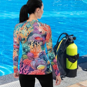 May include: A long-sleeved rash guard featuring a vibrant coral reef design in shades of orange, red, purple, and teal. The shirt is worn by a person standing near a pool with a scuba diving tank.