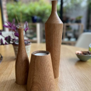 Könnte beinhalten: Drei dekorative Holzobjekte: eine hohe Vase, ein flaschenförmiges Stück und ein konischer Kerzenhalter. Die Objekte sind aus hellbraunem Holz gefertigt und stehen auf einem Holztisch. Der Hintergrund ist verschwommen und zeigt Grün.