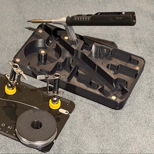 May include: A black soldering station with a built-in magnifying glass and a spool of solder wire. Four helping hands with yellow bases are on a black mat with a ruler. The text "TS101" is visible on the soldering station.  "OmniFixo Helping Hands and TS101 Not Included" is written below the image.