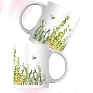 May include: Two white ceramic mugs with a floral and bee design. The mugs feature illustrations of yellow flowers, green foliage, and flying bees. The mugs have a curved handle and a white background.