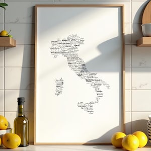 Puede incluir: Un cuadro con un mapa de Italia compuesto por palabras italianas en escritura negra. El cuadro está en un marco de madera clara, expuesto sobre una encimera blanca con limones, una botella de aceite de oliva y una pila de platos blancos.