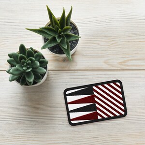 May include: Rectangular patch with a black border, featuring a geometric design. The design includes white, black, and burgundy triangles and diagonal stripes. Two small potted succulents are in the background.