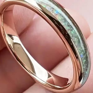 May include: Rose gold ring with a band of iridescent opal. The ring has a smooth, polished finish and a classic, minimalist design. The opal band displays shades of green, blue, and white. This ring is a piece of jewelry.