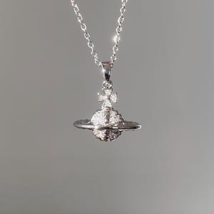 Puede incluir: Collar plateado con un colgante en forma de planeta, adornado con piedras brillantes y una cruz. El colgante cuelga de una delicada cadena plateada. El diseño del planeta es una característica destacada del collar.