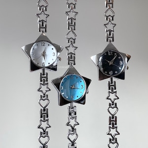 Puede incluir: Tres relojes plateados en forma de estrella con esferas blancas, azules y negras. Cada reloj tiene una cadena con eslabones en forma de estrella y corazón. Las esferas muestran los números 12, 3, 6 y 9.