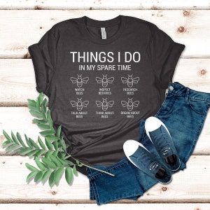 Puede incluir: Una camiseta gris oscuro con el texto "THINGS I DO IN MY SPARE TIME" y una serie de iconos con tem&aacute;tica de abejas. Los iconos representan observar abejas, inspeccionar colmenas, investigar abejas, hablar de abejas, pensar en abejas y so&ntilde;ar con abejas.