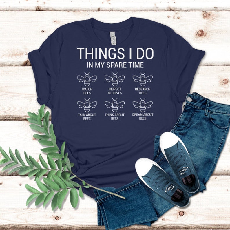 Puede incluir: Una camiseta azul marino con texto blanco que dice "Things I do in my spare time". La camiseta presenta seis ilustraciones de abejas blancas con texto debajo de cada abeja que dice "Watch Bees", "Inspect Beehives", "Research Bees", "Talk About Bees", "Think About Bees", y "Dream About Bees".
