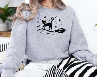 Puede incluir: Sudadera gris claro con un diseño de gato negro sobre una escoba, rodeado de murciélagos y estrellas. El gato tiene ojos amarillos. El diseño está centrado en la parte delantera de la sudadera. La sudadera es de manga larga y tiene cuello redondo.