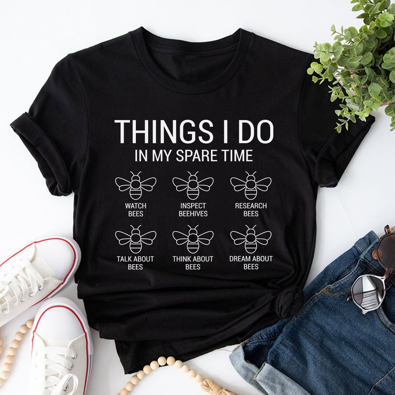 Puede incluir: Camiseta negra con texto blanco que dice "Things I do in my spare time" y seis ilustraciones de abejas con texto que describe actividades relacionadas con las abejas.