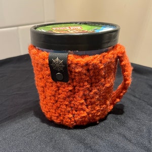 May include: Orange crocheted ice cream container cozy with a black spider web design and a black leather strap.
