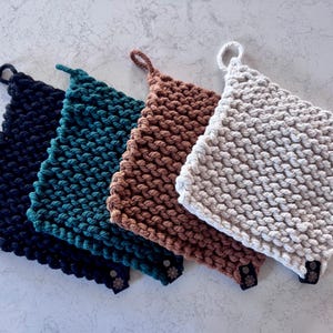 May include: Four handmade knitted pot holders in navy blue, teal, brown, and cream. Each square pot holder has a loop for hanging and a small tag. The pot holders are arranged diagonally, showcasing their textured, chunky knit design.