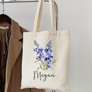 Peut inclure: Un sac fourre-tout en toile blanche avec un motif floral composé de fleurs bleues en forme de cloche. Le sac a une poignée noire et le nom "Megan" est imprimé sur le devant.