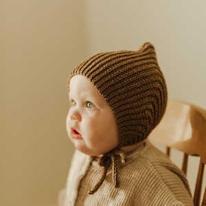 May include: A baby wearing a brown knitted bonnet with a tie, sitting on a wooden chair. The baby is wearing a beige knitted sweater.