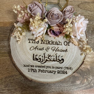 May include: A wooden slice with a decorative floral arrangement and a small glass box. The wood is engraved with the text "The Nikkah Of Arub & Haseeb" in cursive script, followed by Arabic calligraphy and the text "'And we created you in pairs' (78:8) 17th February 2024".