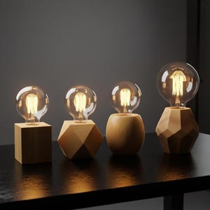 May include: Four decorative wooden table lamps with exposed filament bulbs. The lamps have different geometric wooden bases: a cube, a faceted shape, a rounded shape, and a multi-faceted shape. The bulbs are clear glass and emit a warm, yellow light.