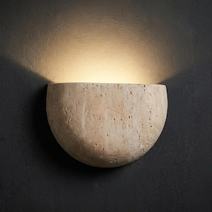 Può includere: Applique da parete beige, a forma di semicerchio, con superficie strutturata. L'applique è montata su una parete grigio scuro e una luce calda brilla dall'interno, creando un bagliore soffuso. Il design è minimalista e moderno.