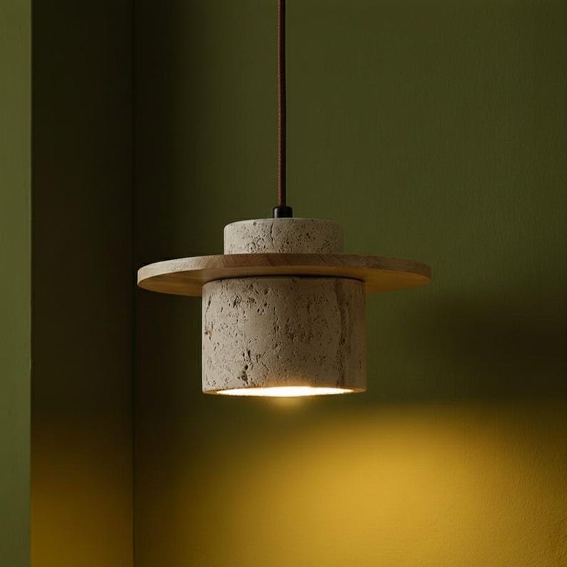 Puede incluir: Una moderna l&aacute;mpara colgante con una pantalla cil&iacute;ndrica de hormig&oacute;n texturizado. Un disco de madera y otro de hormig&oacute;n m&aacute;s peque&ntilde;o est&aacute;n encima de la pantalla. La luz cuelga de un cable marr&oacute;n sobre un fondo verde. La luz est&aacute; encendida.