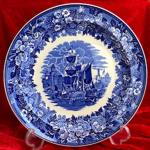 May include: A blue and white decorative plate featuring a nautical scene with ships and buildings. The plate has a floral border in shades of blue. The plate is displayed on a clear stand against a red background.