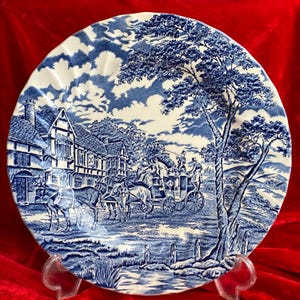 May include: A blue and white plate with a detailed landscape scene. The design shows a horse-drawn carriage, buildings, and trees. The plate has a scalloped edge and is displayed on a clear stand.