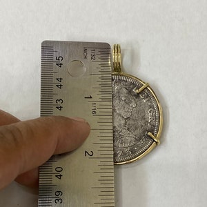 May include: A silver coin with a gold wire wrap and a brass jump ring. The coin is 2 inches in diameter and features a portrait of a man with the text "GR" on the side. The coin is being measured with a ruler.