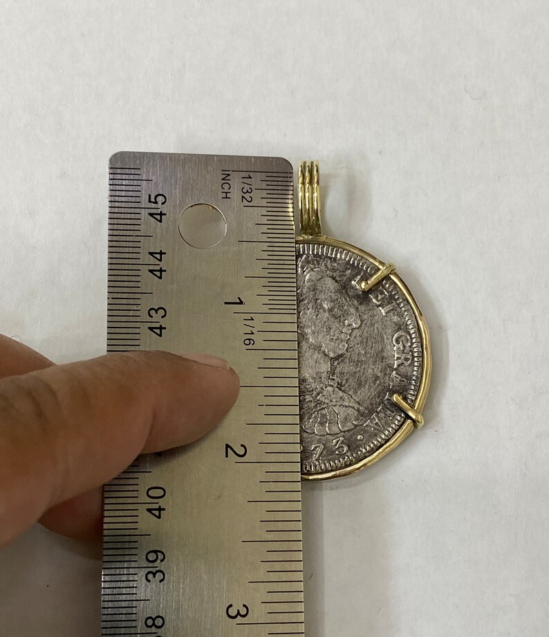 May include: A silver coin with a gold wire wrap and a brass jump ring. The coin is 2 inches in diameter and features a portrait of a man with the text "GR" on the side. The coin is being measured with a ruler.