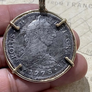 May include: A silver coin with a portrait of a person on one side and the text "REX  GRATIA" on the other side. The coin is dated 1773 and is set in a gold frame.