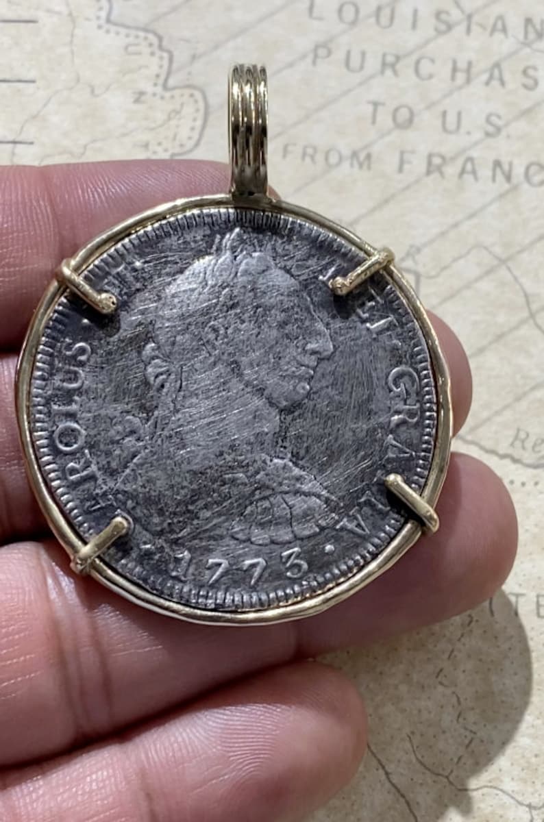 May include: A silver coin with a portrait of a person on one side and the text "REX  GRATIA" on the other side. The coin is dated 1773 and is set in a gold frame.