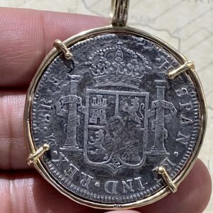 May include: A silver coin pendant with a gold frame. The coin features a coat of arms with a crown and the text "IND. REX. HISPAN."