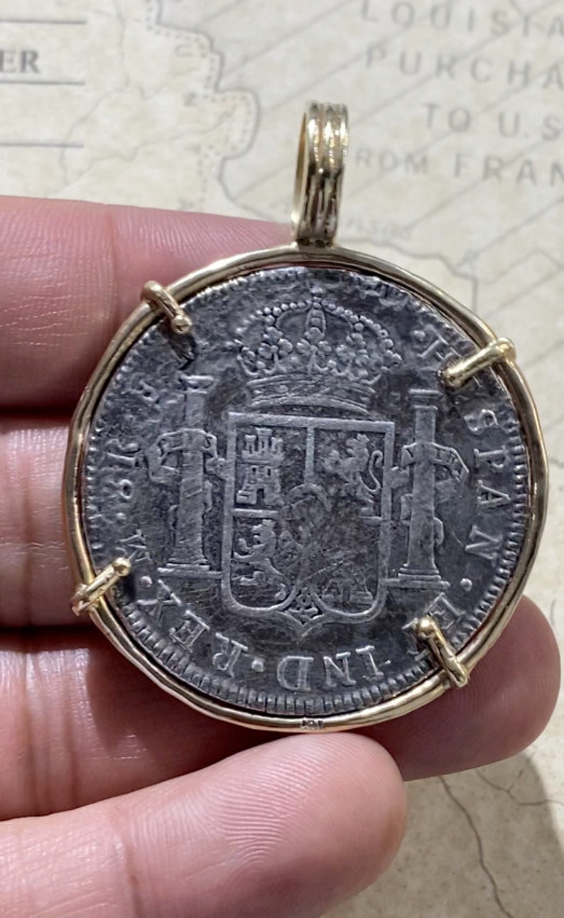 May include: A silver coin pendant with a gold frame. The coin features a coat of arms with a crown and the text "IND. REX. HISPAN."