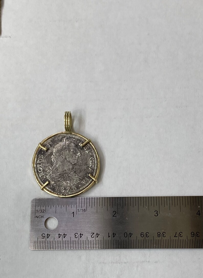 May include: A silver coin with a gold rim and a gold bail. The coin features a portrait of a man with a crown and the text "ROLUS GR" on the edge. The coin is about 1 inch in diameter.