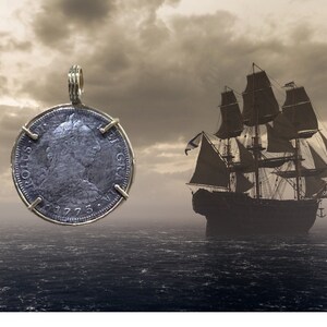 May include: A silver coin pendant with a portrait of a person on one side and the text "ROTUS GR", "1775", and "1773" on the other side. The pendant is hanging from a gold chain against a backdrop of a cloudy sky and a ship sailing on the ocean.