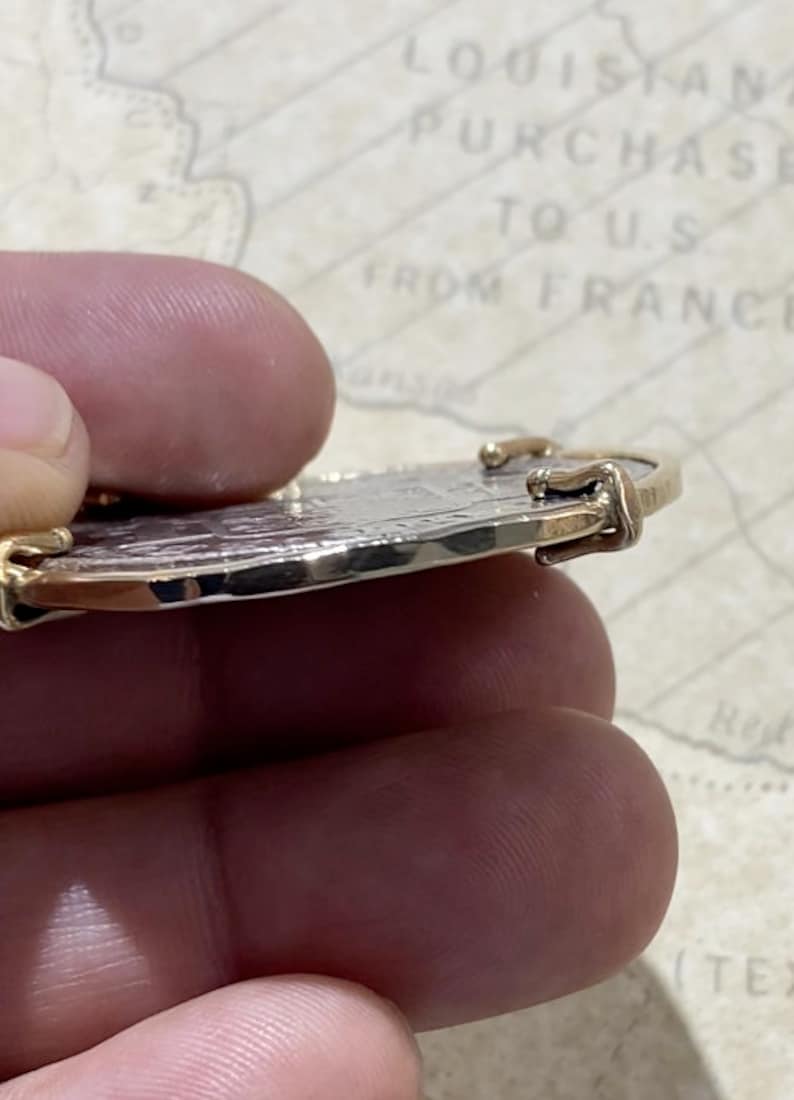 May include: A silver coin with a gold rim and two gold clasps on the side. The coin is being held by a hand.