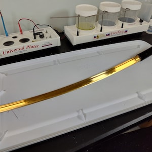 May include: A gold-plated katana sword with a black handle, resting on a white tray. The sword is about 30 inches long and has a slight curve to the blade. The tray is made of plastic and has a textured surface.
