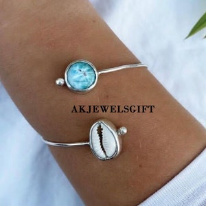Puede incluir: Dos pulseras de plata con un dije de concha azul y blanco y un dije de concha blanca. Las pulseras están en la muñeca de una persona. AKJEWELSGIFT está escrito en la imagen.