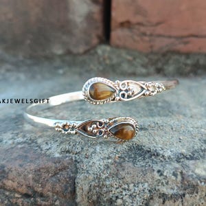 May include: Silver bangle bracelet with two tiger's eye stones. The bracelet is open and has a delicate design.
