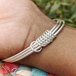 May include: A silver-colored bangle bracelet with a unique design. The bracelet features three parallel bands held together by a series of coiled wire and a circular accent. The text "AKJEWELSGIFT" is visible on the bracelet.