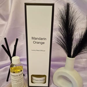 May include: A black box with a white label that reads "Mandarin Orange Luxury Reed Diffuser". A clear glass bottle with a yellow liquid and a white label that reads "Mandarin Orange Luxury Reed & Cane Diffuser Refill" is in front of the box. A white ceramic vase with a black feather plume is on the right side of the image.
