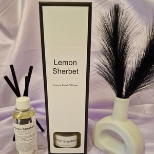 May include: A black box with white text that reads "Lemon Sherbet Luxury Reed Diffuser". A clear glass bottle with a yellow liquid and a white label that reads "Lemon Sherbet Luxury Reed & Car Diffuser Refill" is in front of the box. A black and white vase with a black feather-like plume is on the right side of the image.