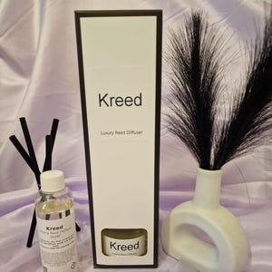 May include: A white bottle with a black label that says "Kreed Luxury Reed Diffuser Refill" is next to a black box with the same text. The box is next to a white ceramic vase with a black feather plume.