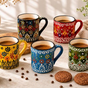 May include: A set of five ceramic mugs featuring intricate, colorful floral patterns in black, red, yellow, blue, and green. Each mug has a matching handle and a white interior, filled with dark coffee, arranged on a light surface with coffee beans.