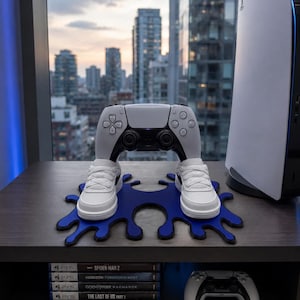 May include: A white and black wireless game controller resting on two miniature white sneakers, which are placed on a blue ink-blot shaped stand. A white game console stands to the right, and five game cases are stacked on the shelf below.
