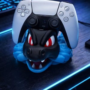 Może przedstawiać: Biało-czarny kontroler PlayStation 5 spoczywa na stojaku na kontroler o tematyce smoka. Stojak jest czarny z czerwonymi oczami i niebieską, falistą podstawą. Kontroler jest biały z czarnymi akcentami.