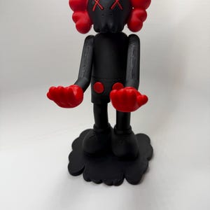 Puede incluir: Una figura negra con detalles rojos. La figura tiene una cabeza con forma de calavera con ojos tachados, manos rojas y protuberancias rojas en forma de oreja. Se apoya sobre una base negra en forma de nube. El cuerpo es negro con detalles circulares rojos.